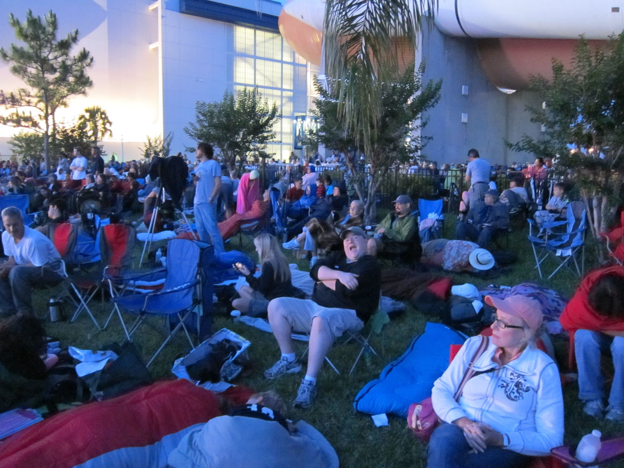 Large crowd of spectators on the lawn at Kennedy Space Center waiting for the shuttle launch at dawn, with rocket exhibit visible overhead