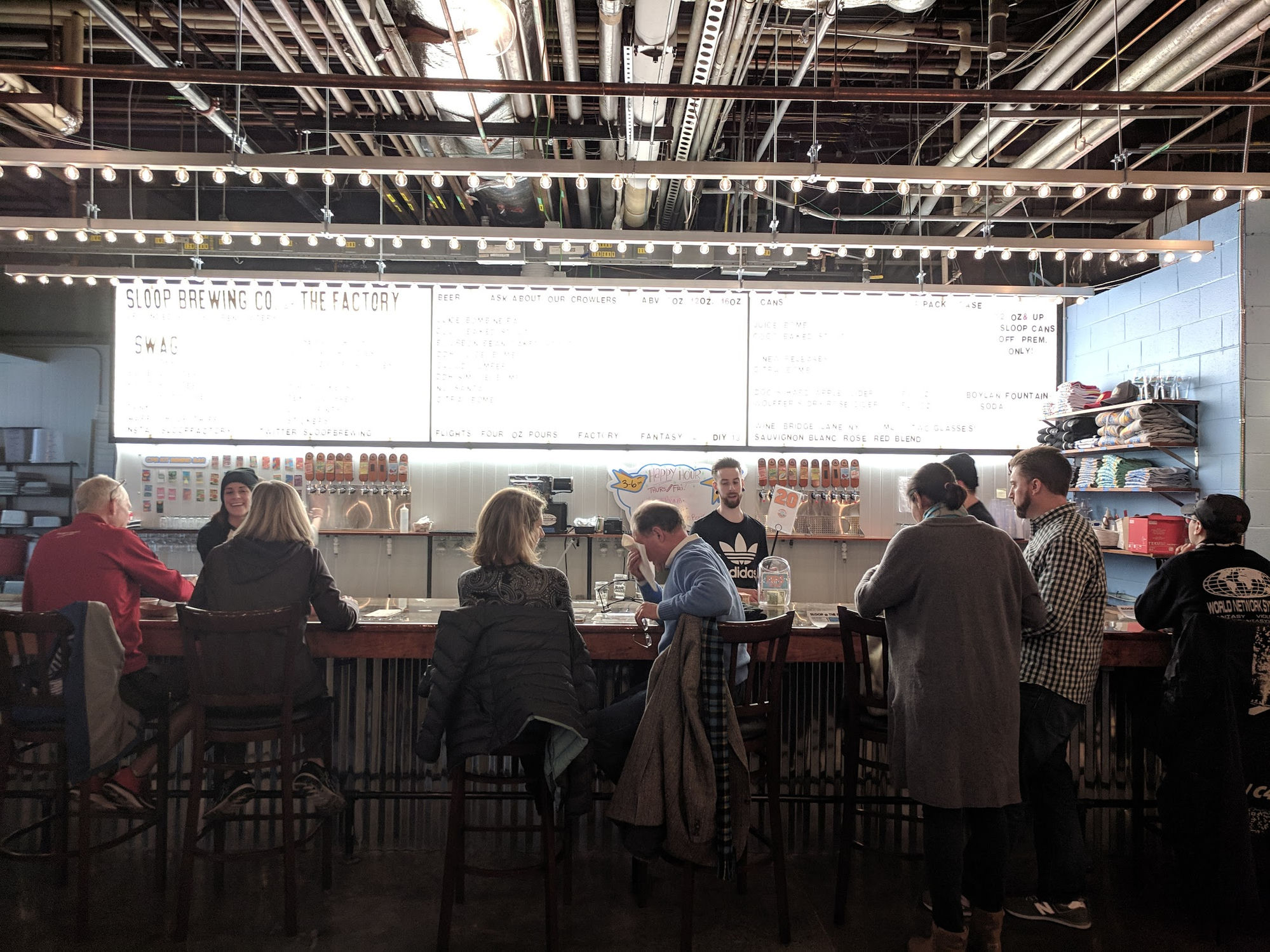 The bar at Sloop Brewing Co. The Factory — patrons seated along a long bar with the full tap and menu board behind