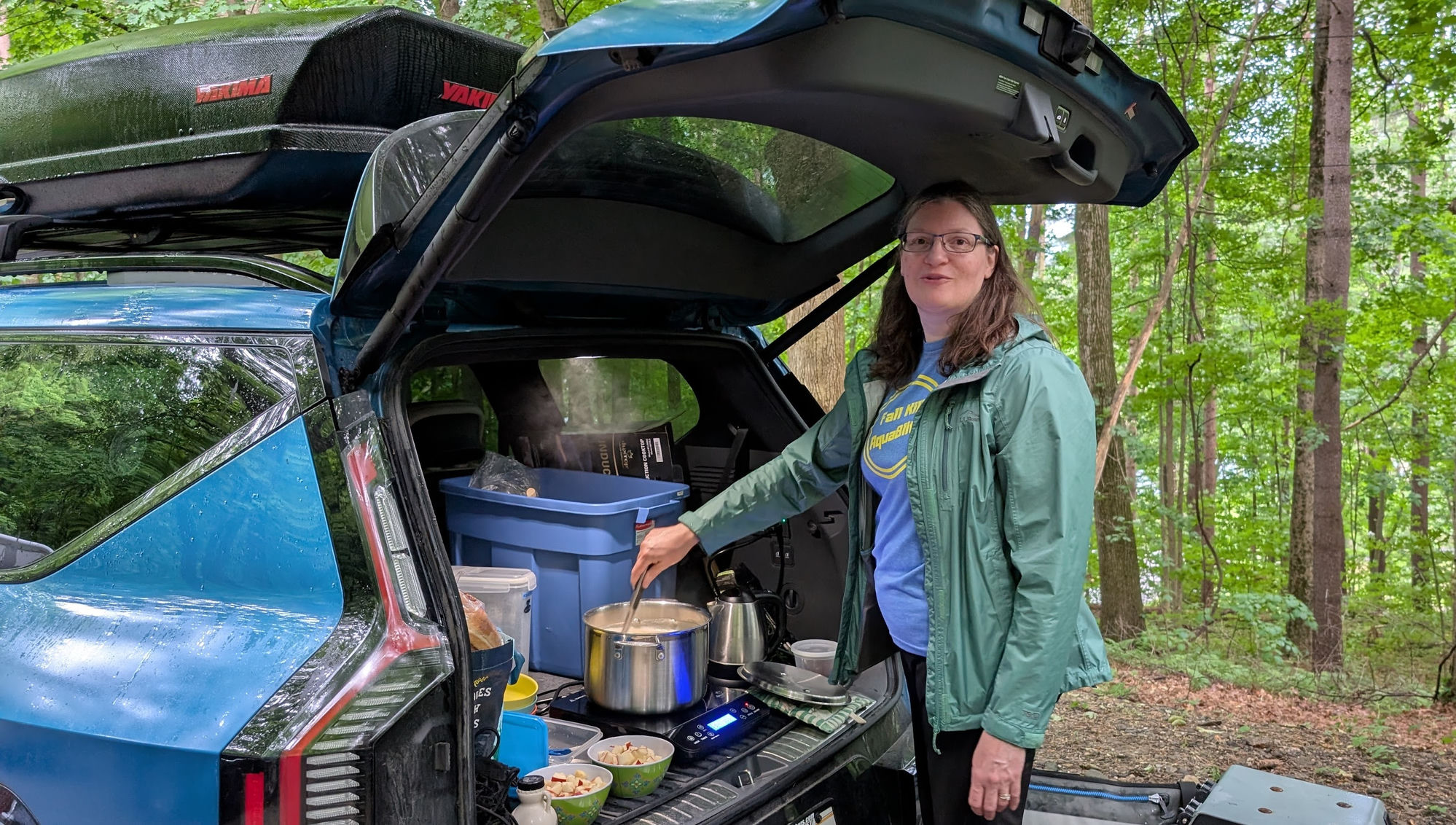 Person cooking on an induction hotplate in the open tailgate of a blue Kia EV9 at a wooded campsite.