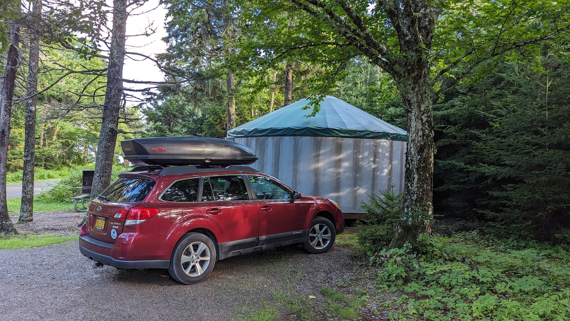 Red Subaru Outback with roof cargo box parked in front of a yurt at a wooded campsite